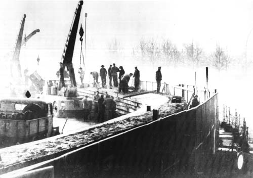 Construction of the Berlin Wall: a misty black and white image of work on the wall, cranes and workers left, the shadow of the no-man's-land area to the right.